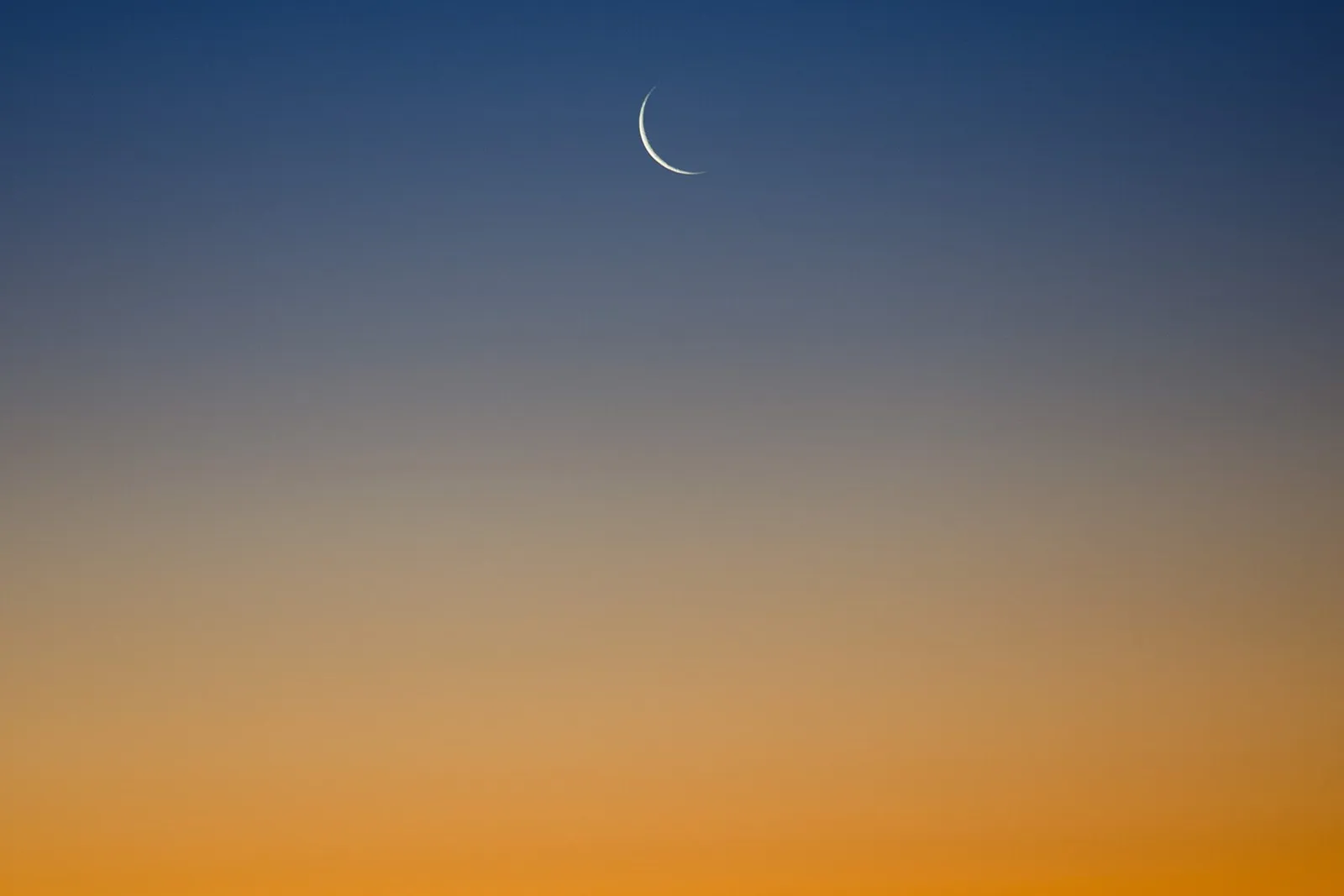 A thin crescent moon in a clear twilight sky fading from deep blue to orange