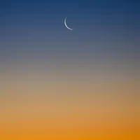 A thin crescent moon in a clear twilight sky fading from deep blue to orange.