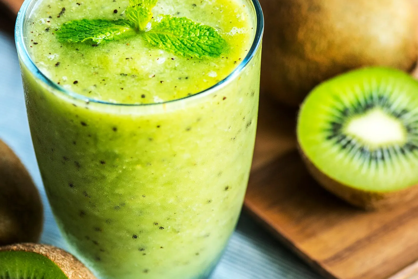 A glass of green kiwi smoothie topped with mint, beside halved kiwi fruit on a wooden board