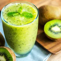 A glass of green kiwi smoothie topped with mint, beside halved kiwi fruit on a wooden board.