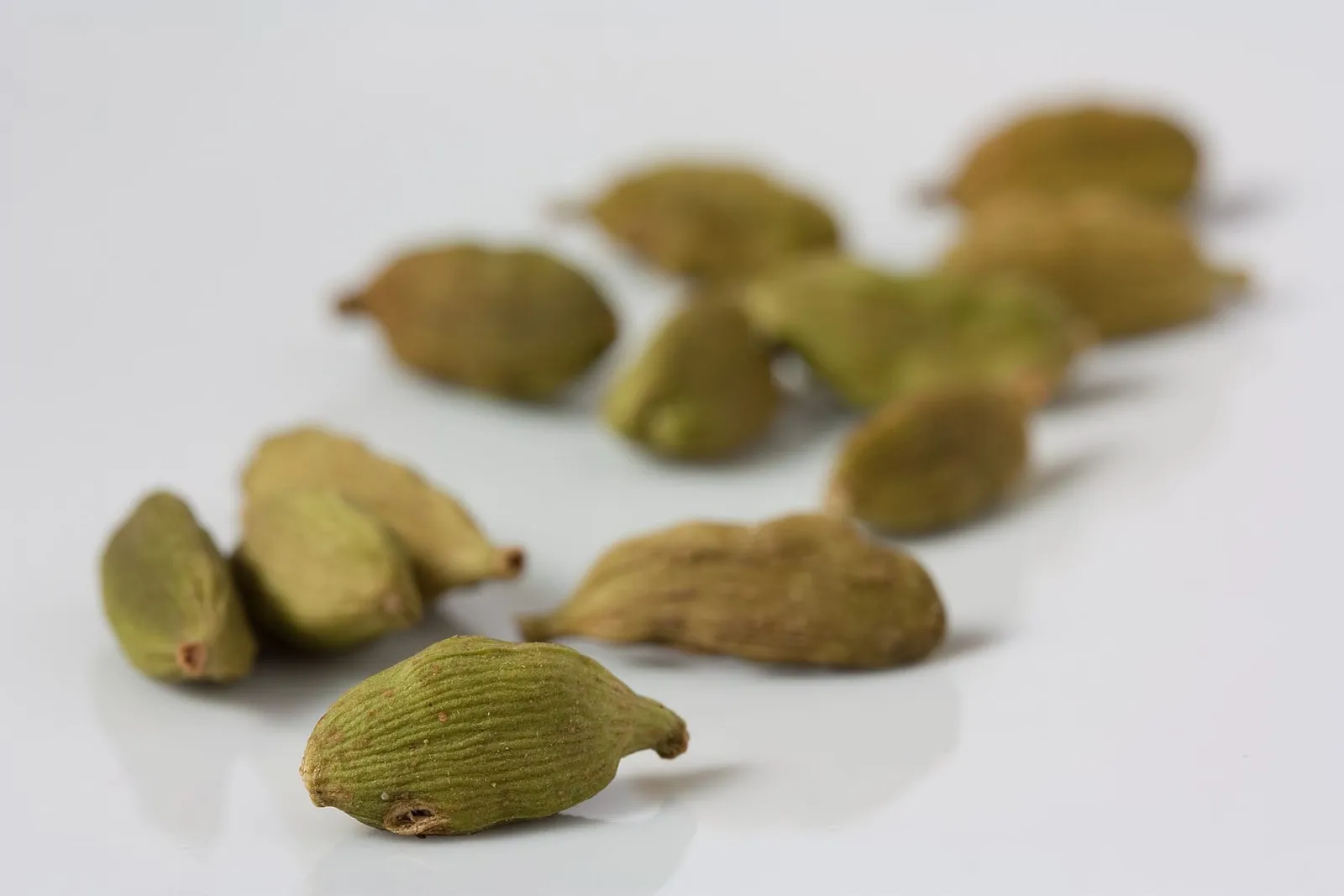 Dried green cardamom pods with fine ridges scattered across a pale white surface.