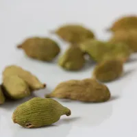 Dried green cardamom pods with fine ridges scattered across a pale white surface.