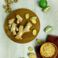 Fresh ginger roots and slices on a round wooden board, with limes and a bowl of ground ginger on a pale table.