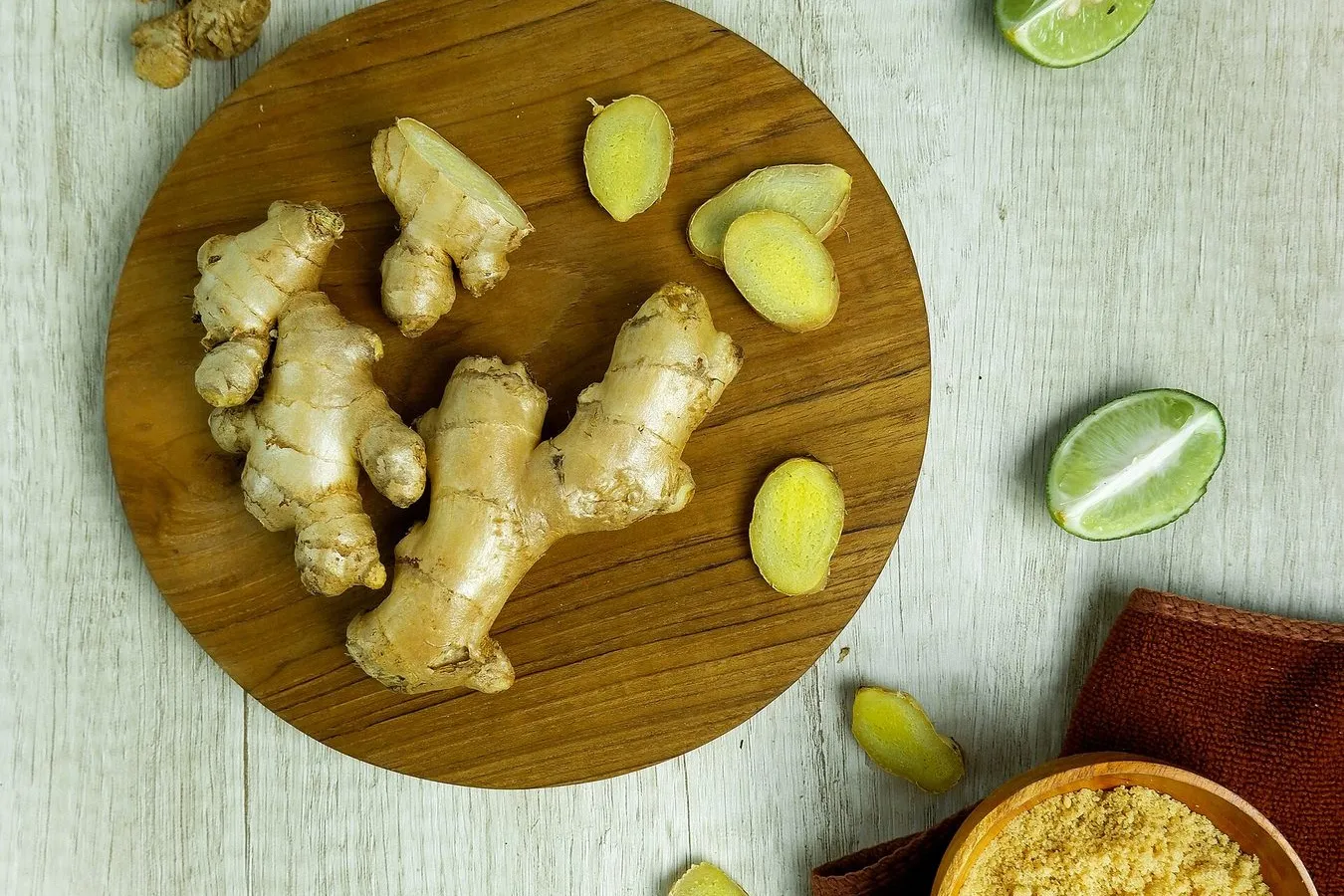 Fresh ginger roots and slices on a round wooden board, with limes and a bowl of ground ginger on a pale table.