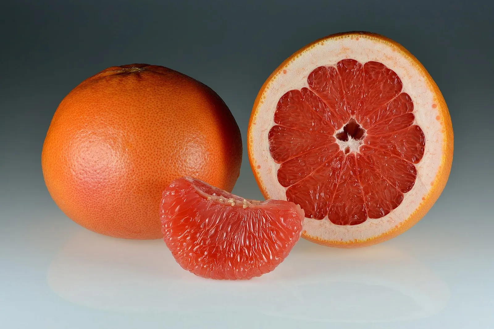 A whole red grapefruit beside a halved one and a single peeled segment on a pale background.