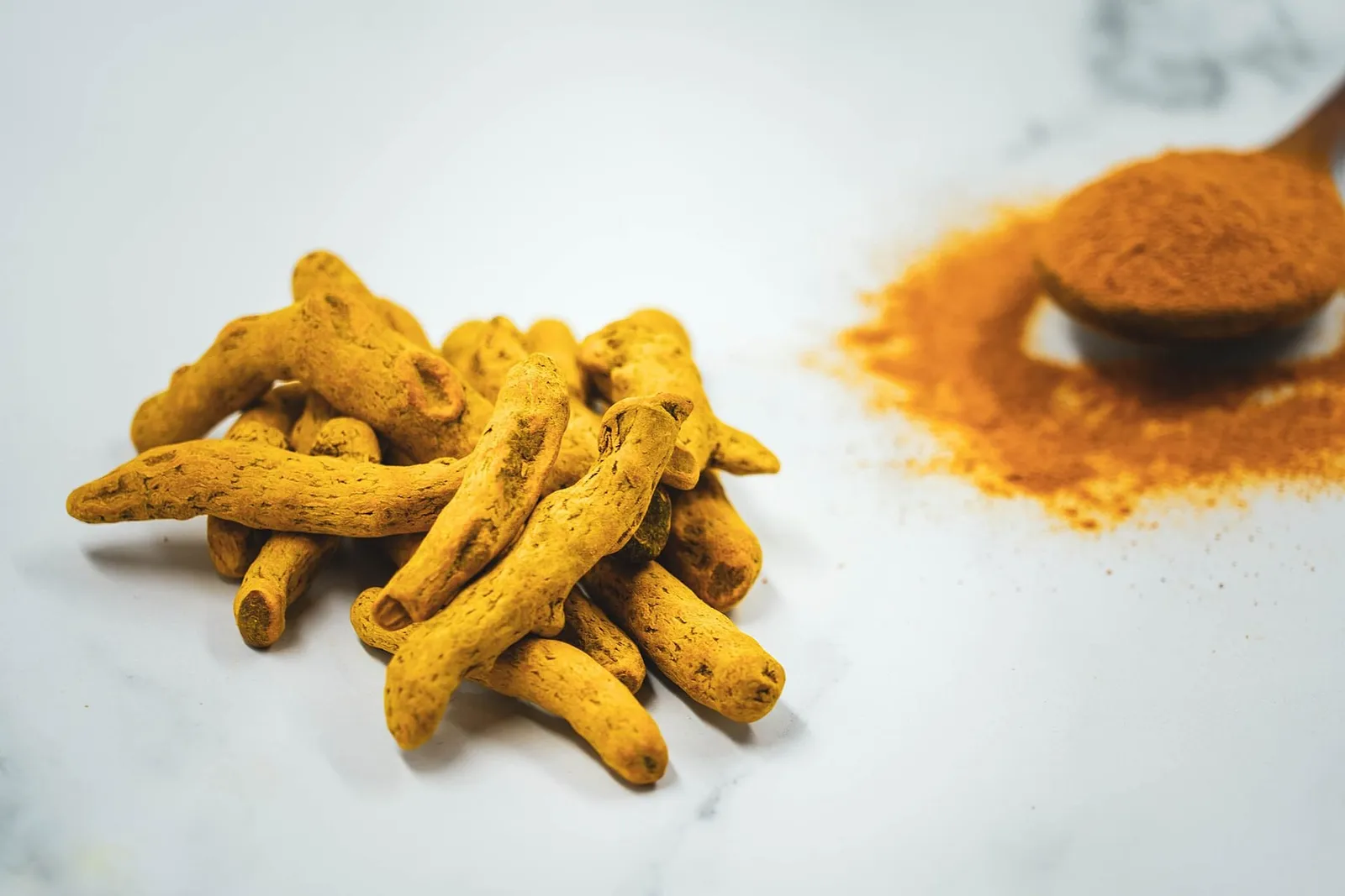 A pile of dried golden-yellow turmeric rhizomes on a white marble surface beside a spoon of ground turmeric.