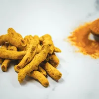 A pile of dried golden-yellow turmeric rhizomes on a white marble surface beside a spoon of ground turmeric.