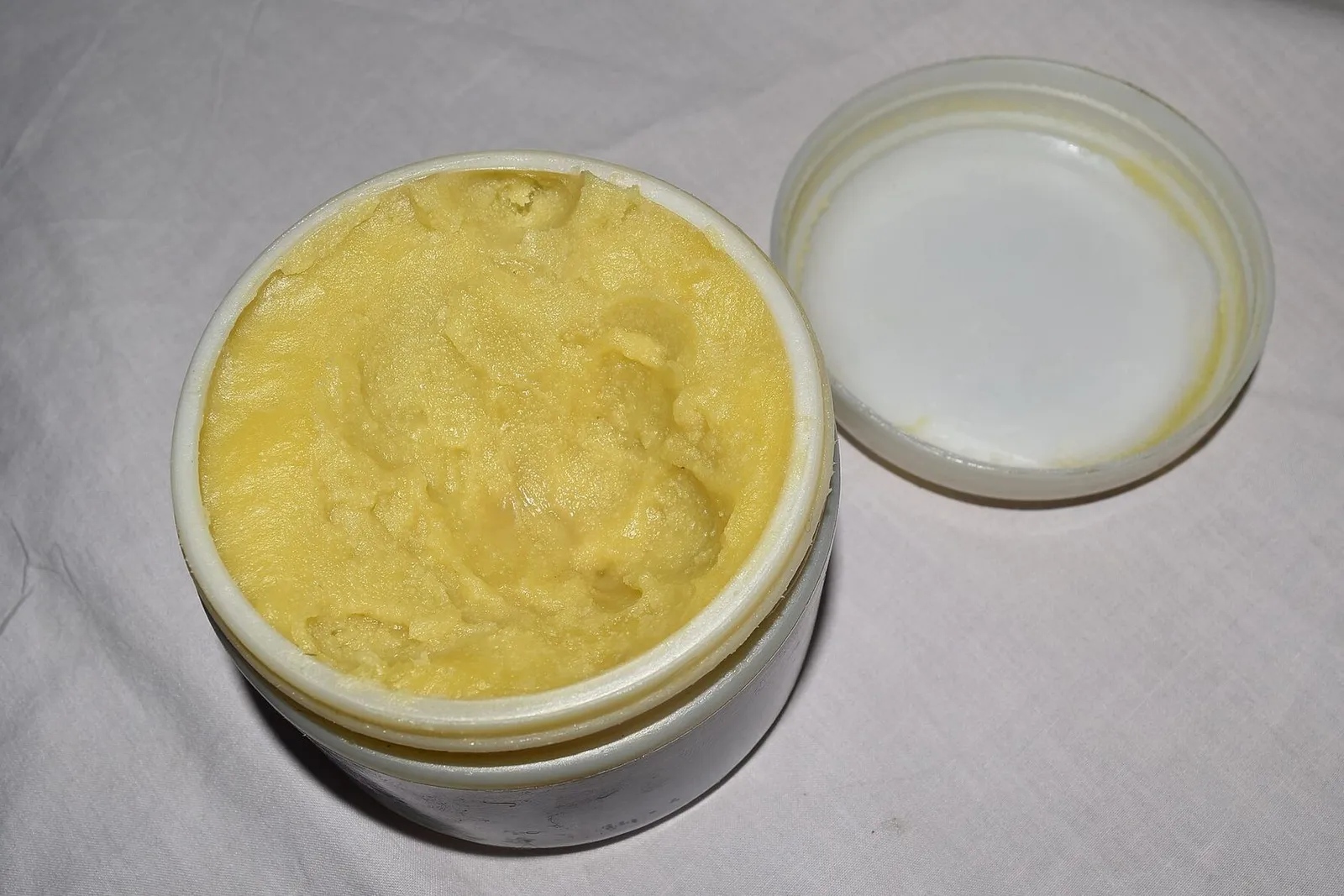 An open tub of whipped shea butter blended with essential oils, its lid set beside it on a pale cloth