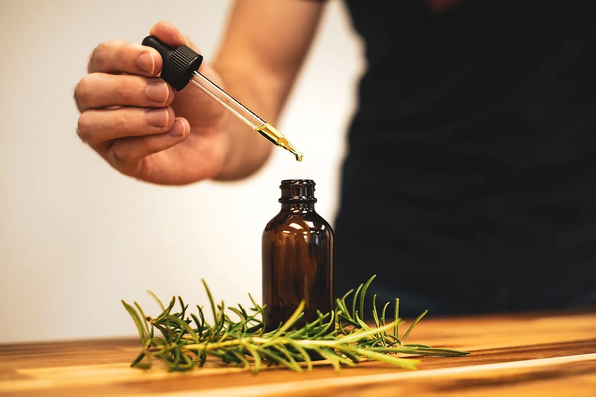 A hand lifting a glass dropper out of a small amber bottle, with rosemary sprigs on the table below.