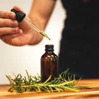 A hand lifting a glass dropper out of a small amber bottle, with rosemary sprigs on the table below.