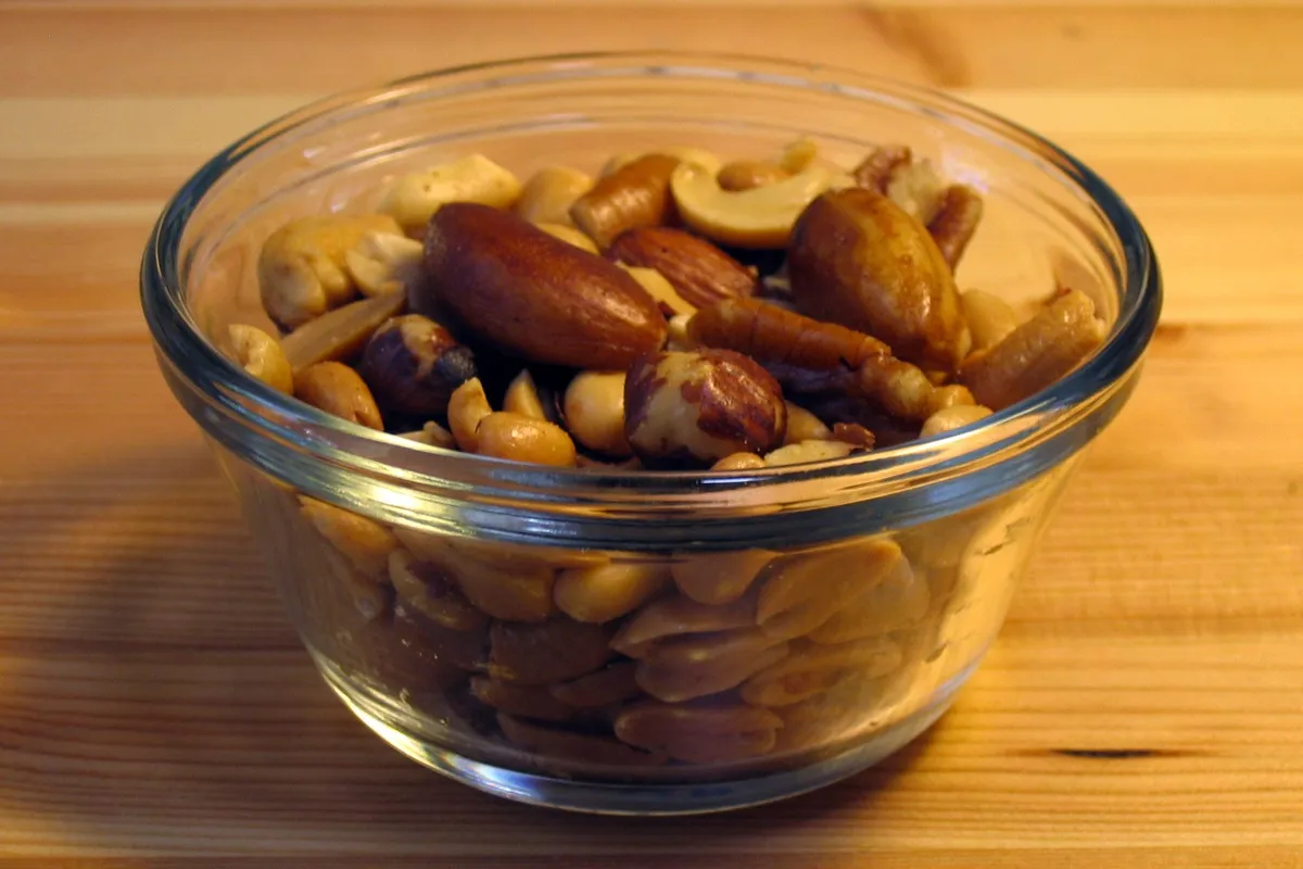 A small glass bowl of mixed nuts, with almonds, Brazil nuts and cashews on top of peanuts, on a wooden table.