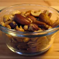 A small glass bowl of mixed nuts, with almonds, Brazil nuts and cashews on top of peanuts, on a wooden table.